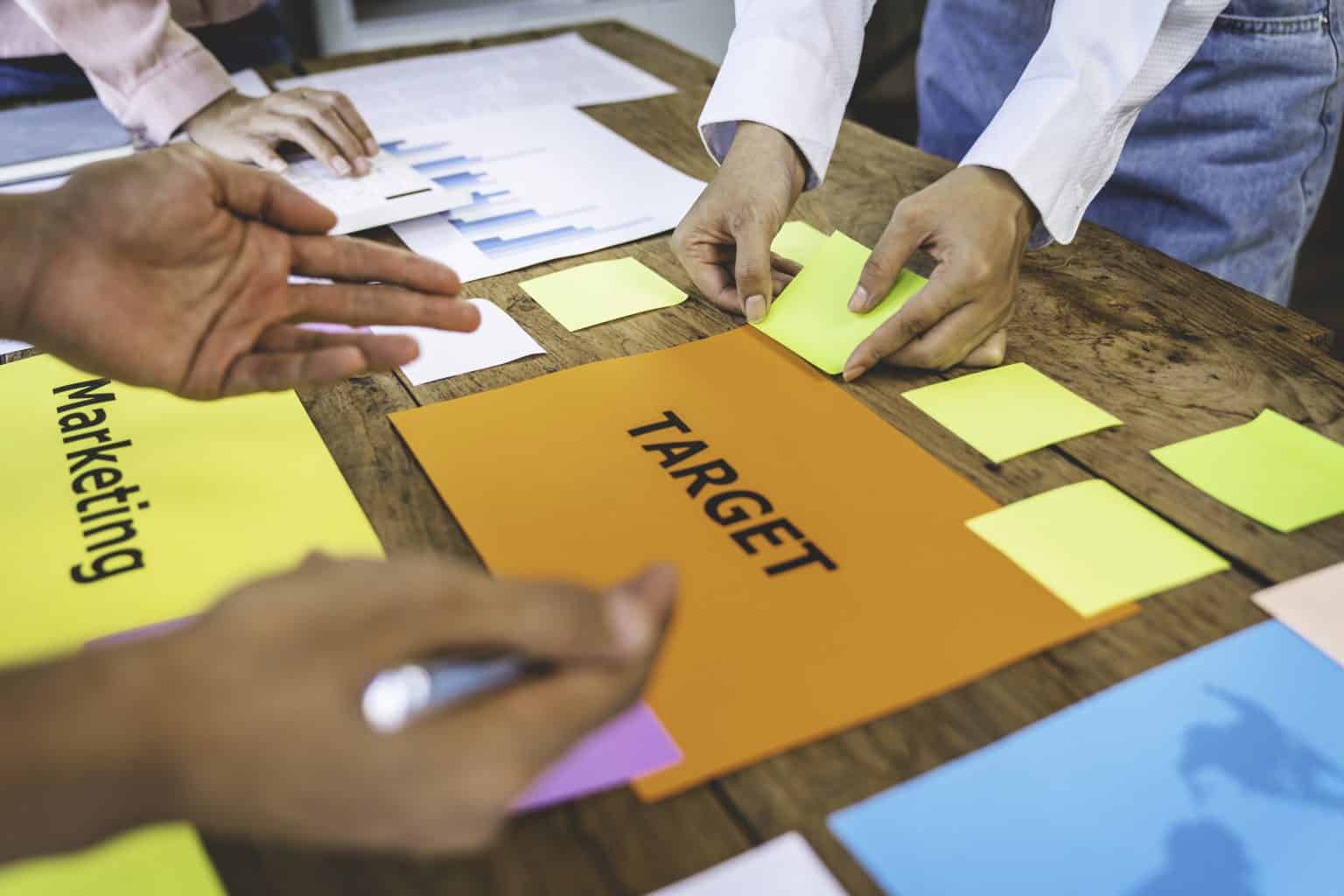 Hands collaborating over a wooden table with colorful sticky notes and papers, focusing on an orange sheet labeled "TARGET," illustrating teamwork in developing marketing strategies for lead generation.