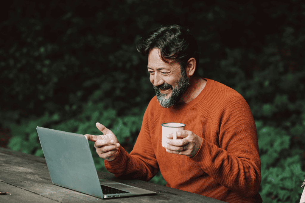 Man in an orange sweater smiling while holding a cup and engaging with a laptop, symbolizing remote work and digital marketing strategies for local business growth.