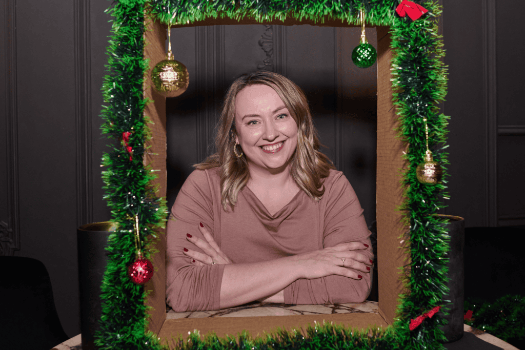 Smiling woman posing inside a festive frame adorned with green garland and ornaments, representing a cheerful holiday atmosphere.