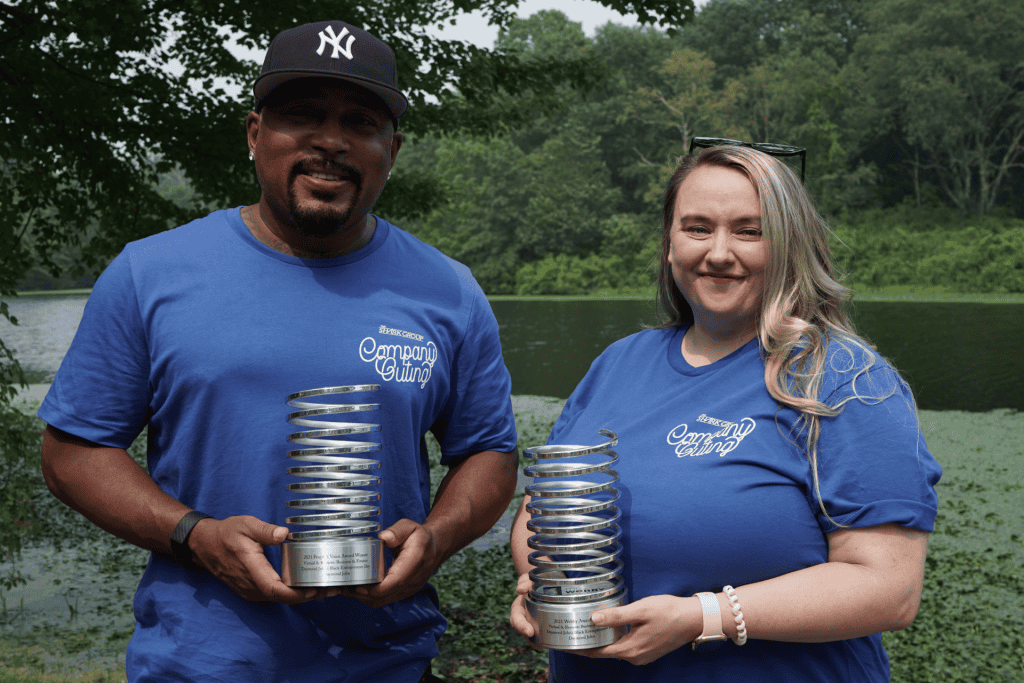 Two individuals holding awards, wearing matching blue shirts with "Company Outing" text, standing by a lake surrounded by greenery, celebrating business achievements.