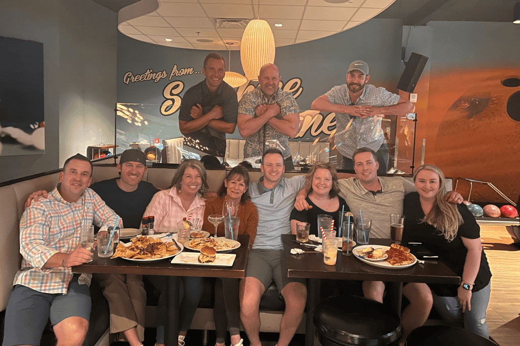 Group of friends enjoying a meal at a bowling alley, featuring various dishes like nachos and burgers, with a vibrant "Greetings from ..." mural in the background.
